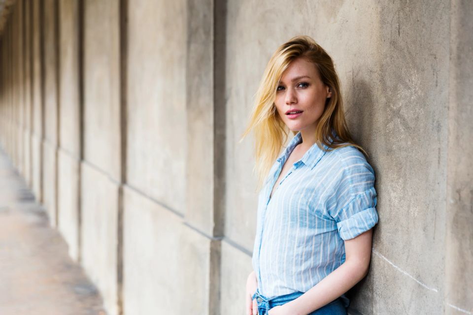 Portrætfoto af ung blond kvinde i lyseblå skjorte, der læner sig afslappet op ad en grå betonvæg med blødt lys.