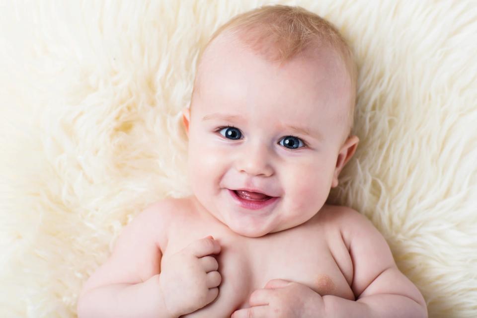 Legende baby fotografering. En smilende baby med lyseblond hår og store blå øjne ligger glad på et lunt fåreskind.