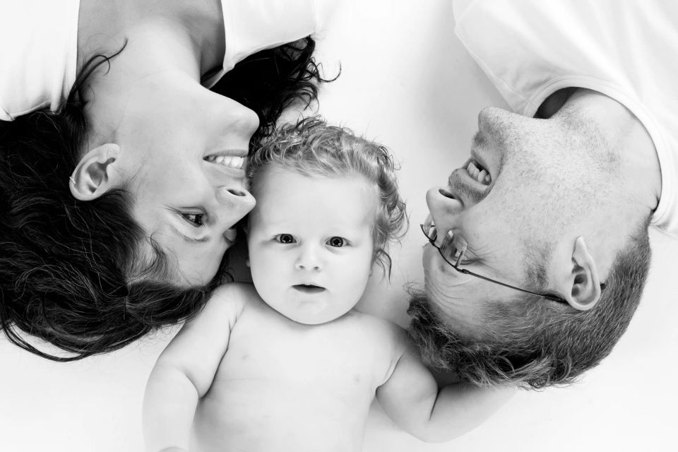 Sort/hvidt babyfotografi af lykkelige forældre, der smiler til deres baby, der ligger i midten på en hvid baggrund.