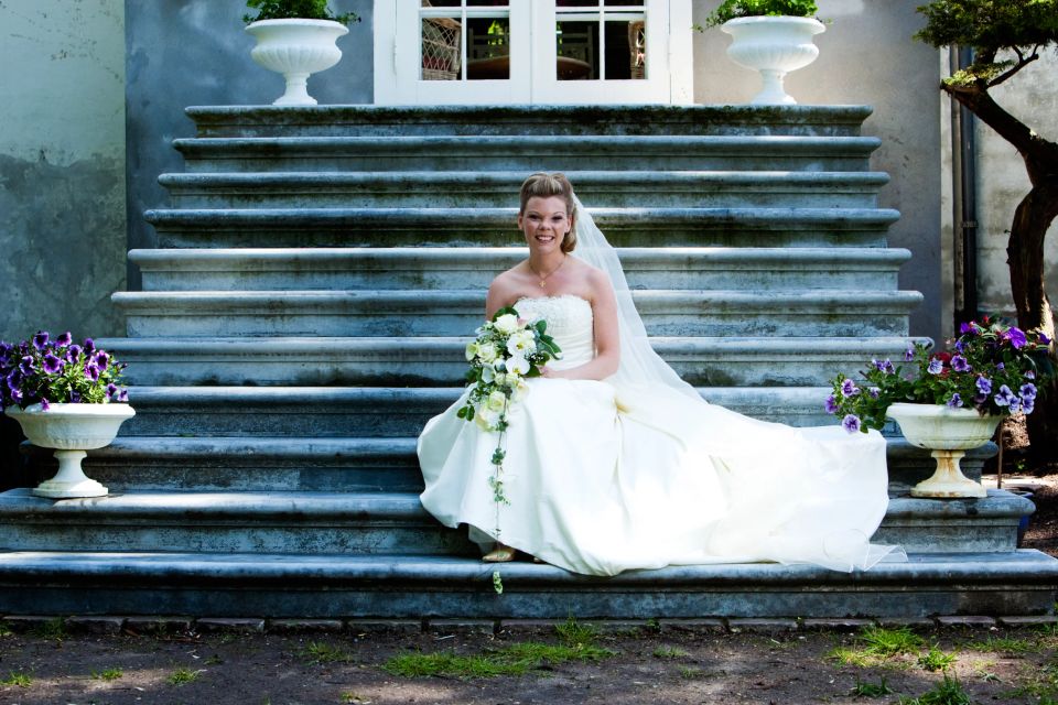 Brud i hvid kjole sidder smilende med brudebuket på gammel stentappe med blomsterkrukker. Bryllupsfotografering.