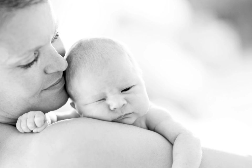 Sort/hvidt babyfotografi af mor der kysser sit spædbarn på hovedet, set fra siden i naturligt, blødt lys.