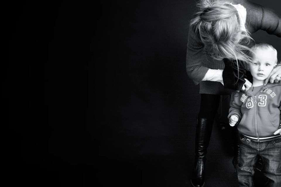 Børneportræt i sort/hvid af en mor med bøjet hoved, der ømt holder om sin søn mod en mørk baggrund.