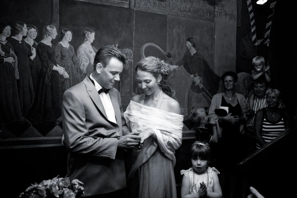 Bryllupsfotografering: Brudgom sætter ring på brudens finger i et rådhus med historiske vægmalerier i baggrunden.