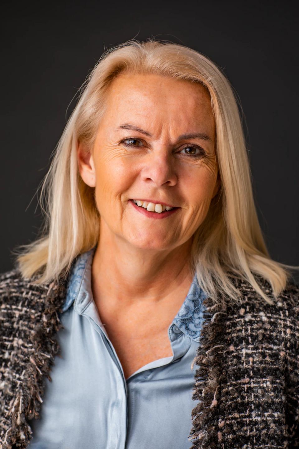Portrætfoto af smilende kvinde med blondt hår i Virum, iført tweedjakke og blå skjorte mod mørk baggrund.