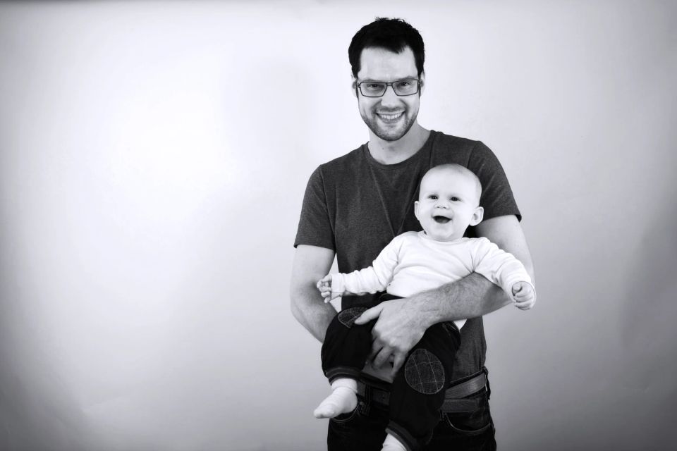 Babyfotografering i sort/hvid af smilende far med briller, der holder glad baby med åben mund og lyse øjne.