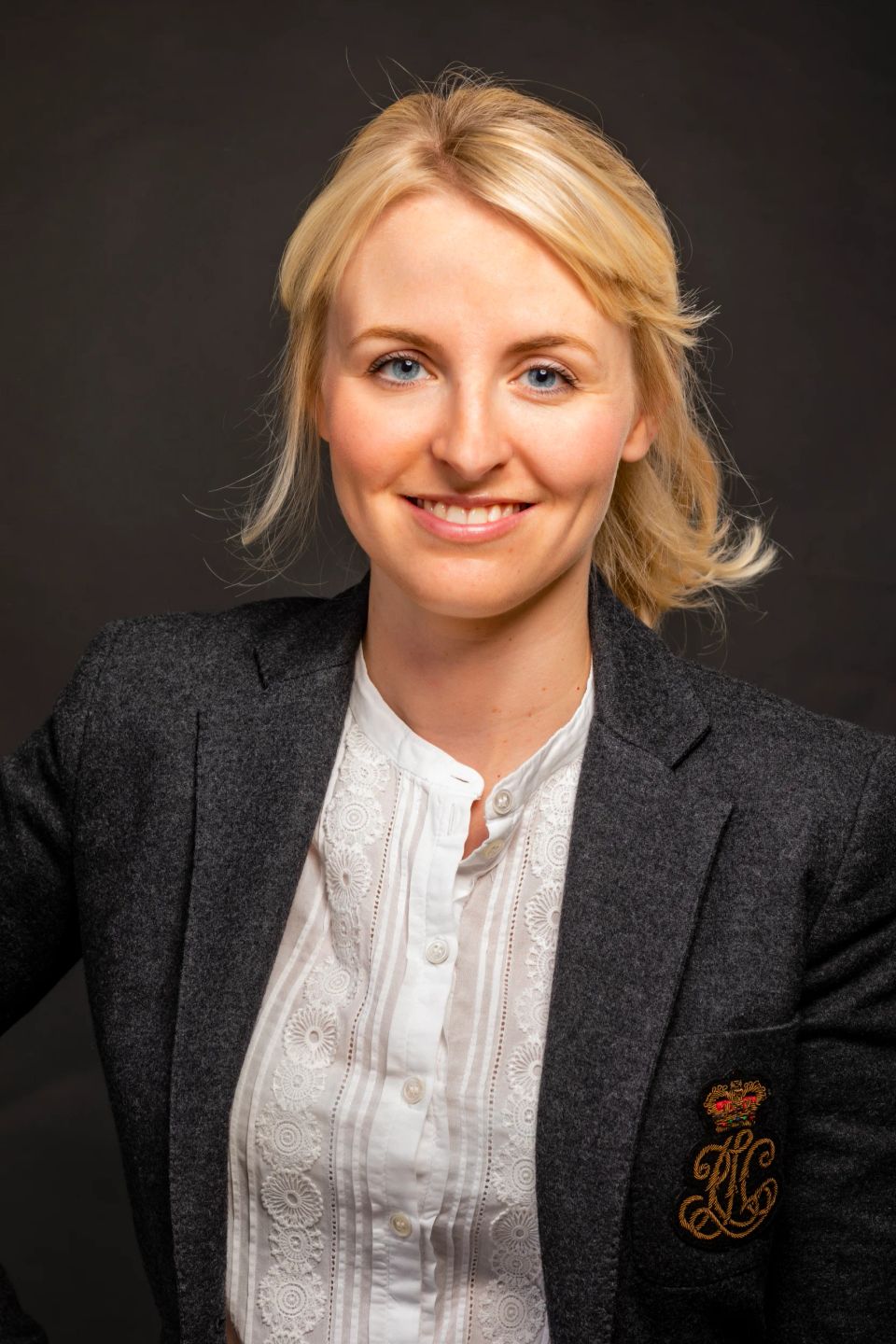 Portrætfoto af en blond kvinde med et autentisk smil, klædt i grå jakke og hvid skjorte med broderi.