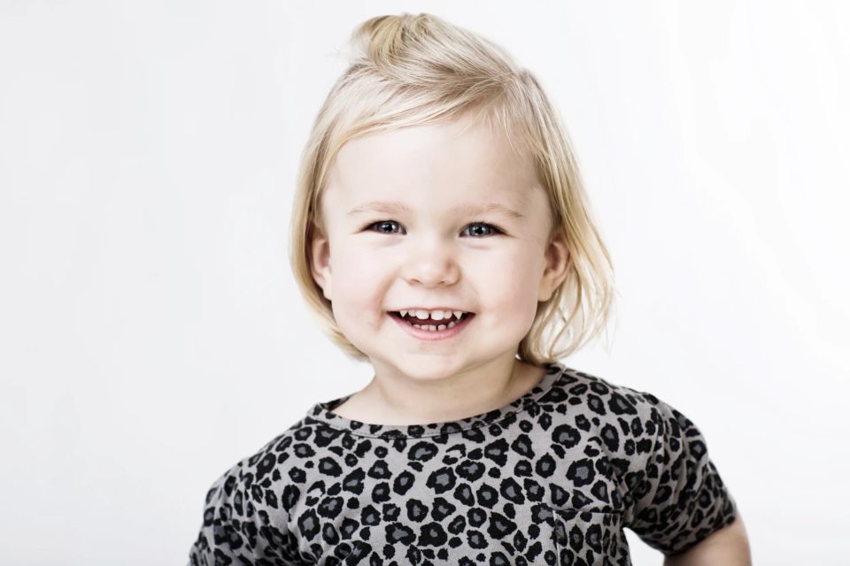 Børnefotografi af en smilende lille pige med lyst hår og leopardprint bluse mod hvid baggrund i blødt lys.