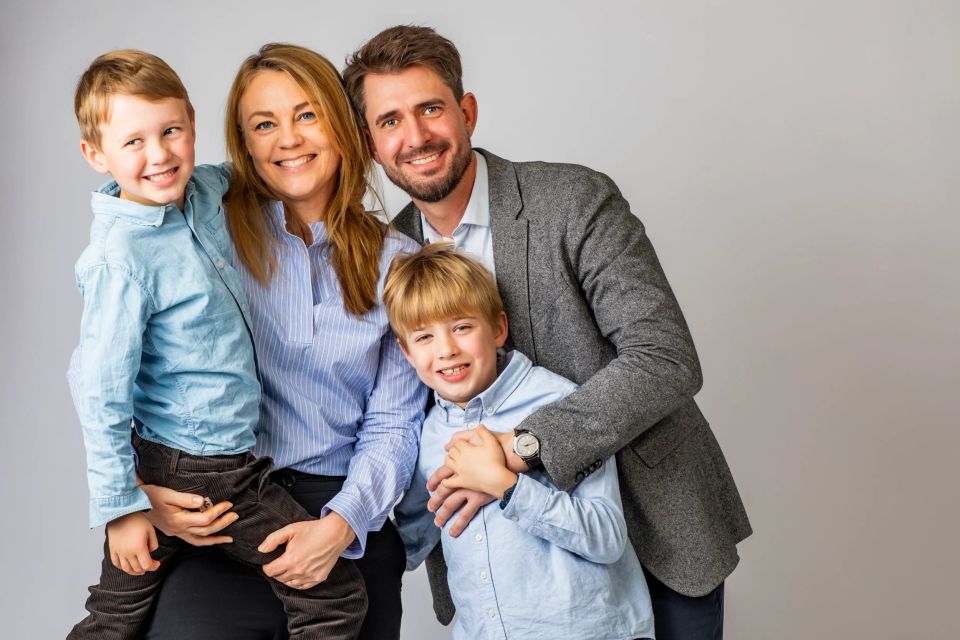 Familiefoto med mor, far og to glade drenge smilende mod kameraet i flot lys og neutral baggrund.