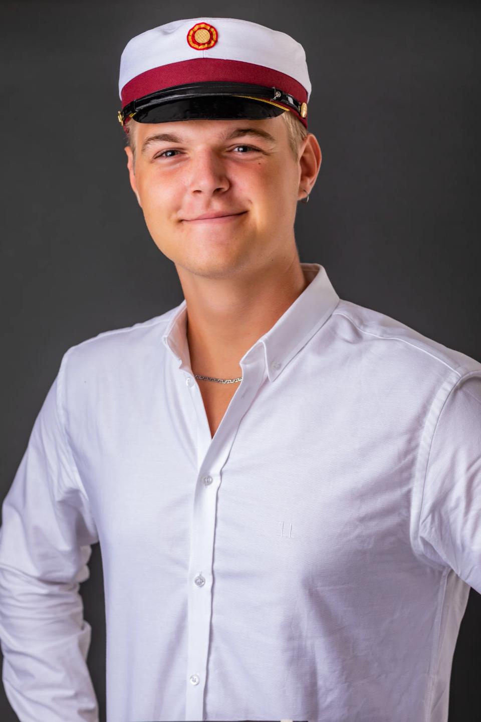Portrætfoto af ung smilende student med hvid studenterhue, hvid skjorte og sølvkæde mod mørk baggrund.