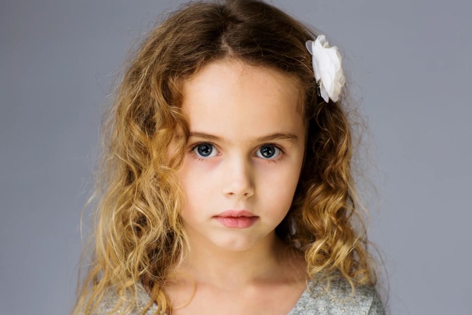 Børnefotografering af pige med krøllet hår, klar blå øjne og hvid blomst i håret mod grå baggrund.