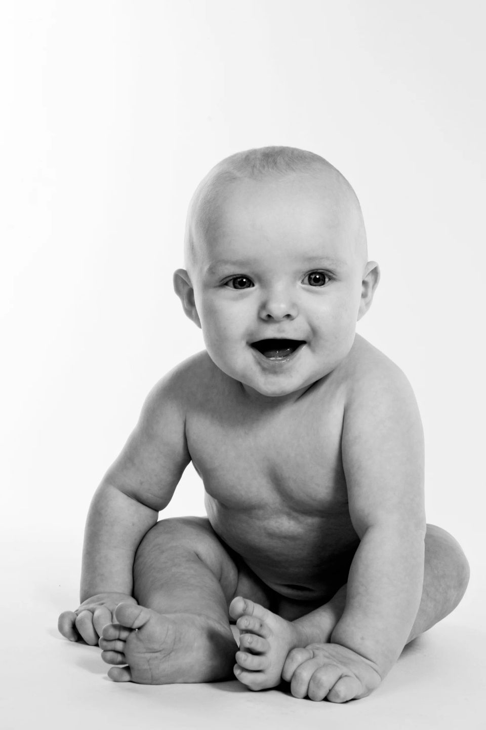 Glad baby sidder med lyst, autentisk smil i sort/hvidt babyfoto. Naturlig lyssætning og neutral baggrund.