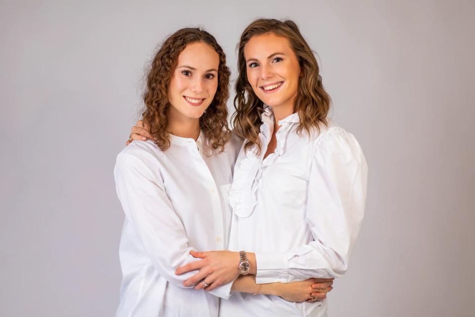 Familieportræt af to smilende søstre i hvide skjorter, der omfavner hinanden, med varme i øjnene.