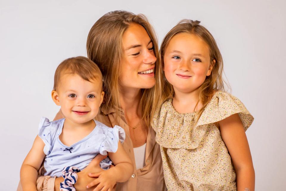 Glad mor med lyst hår holder glad baby, mens ældre pige smiler ved siden af. Et dejligt børnefotografi.