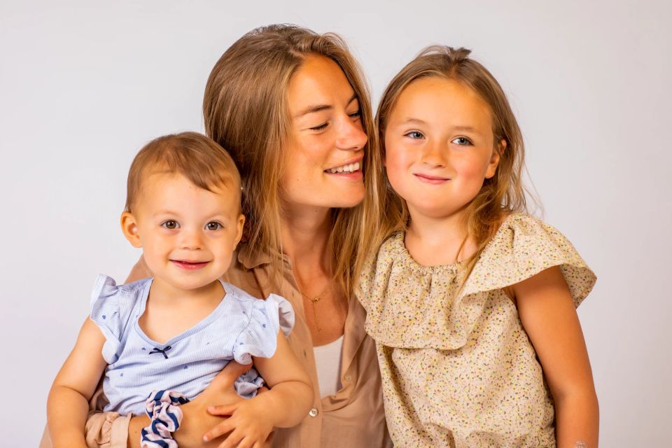 Glad mor med lyst hår holder glad baby, mens ældre pige smiler ved siden af. Et dejligt børnefotografi.