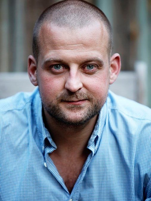 Portrætfoto af mand med blåt blik og skægstubbe i lyseblå skjorte, der ser direkte ind i kameraet.