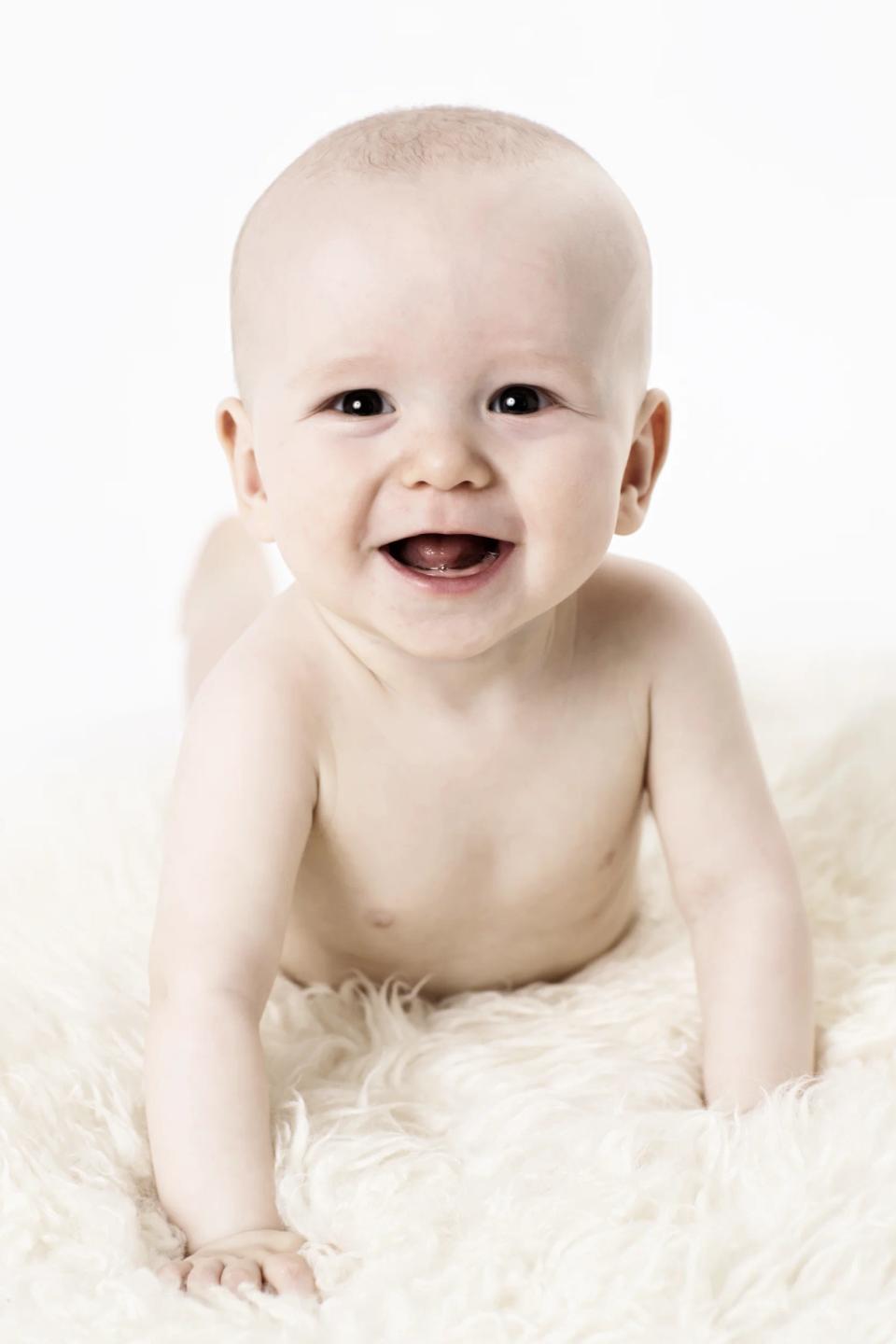 Glad baby ligger på maven på et hvidt, flosset tæppe med et stort smil. Et hjertevarmt babymoment.