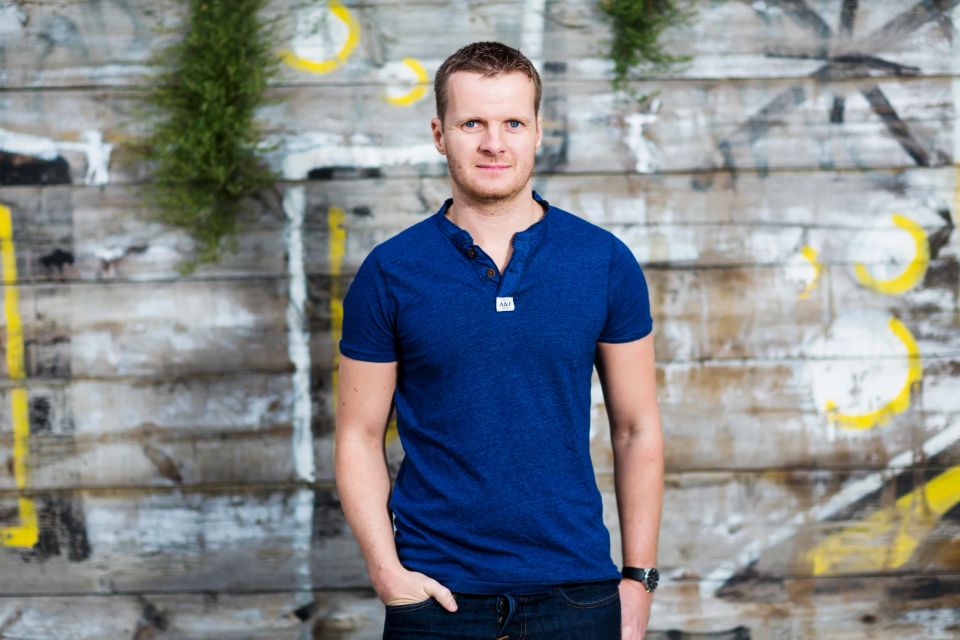 Portrætfoto af smilende mand i blå t-shirt foran rustik trævæg med grønne planter og gult graffiti i Nordhavn.