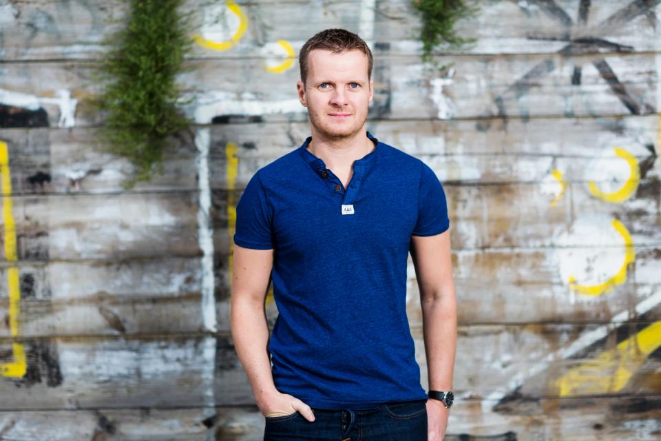 Portrætfoto af smilende mand i blå t-shirt foran rustik trævæg med grønne planter og gult graffiti i Nordhavn.