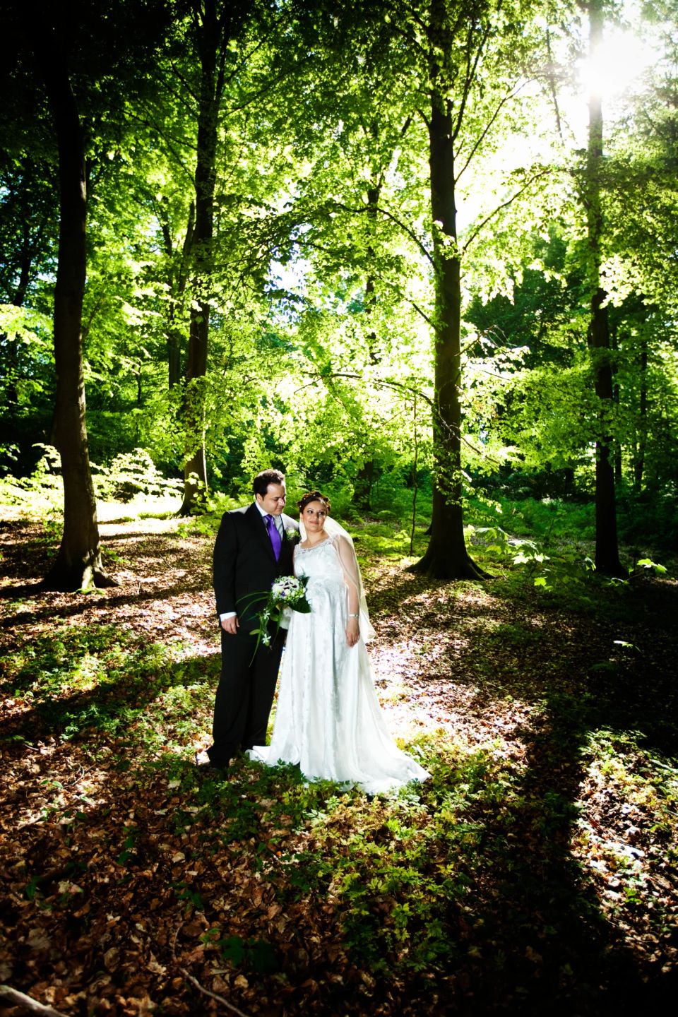 Bryllupsfotografering af smilende brudepar i skov. Sollys filtreres gennem grønne blade og skaber et naturligt look.