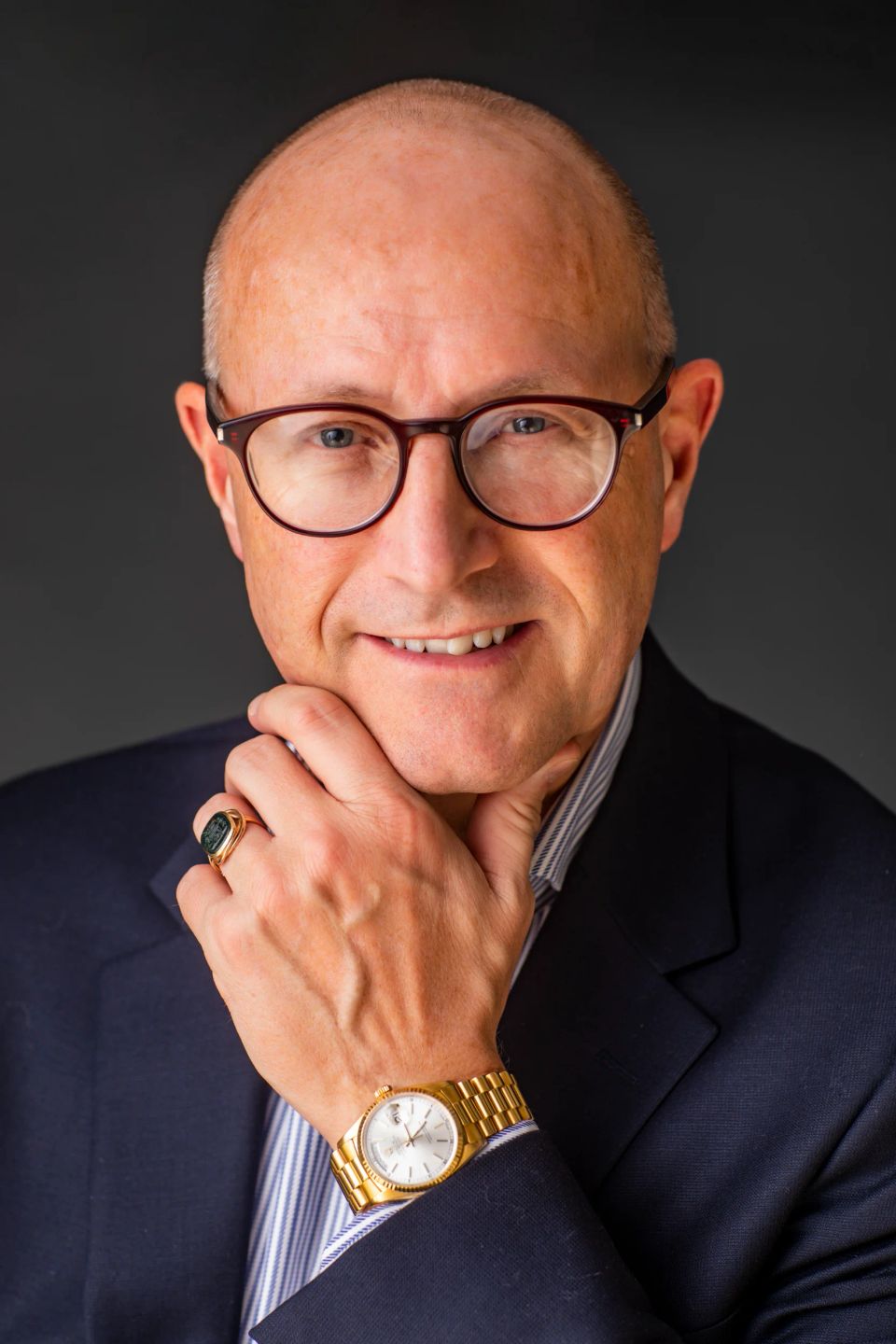 Portrætfoto af smilende, midaldrende mand med briller og guldur, iklædt mørkeblå habitjakke. Han holder sin hånd til hagen.