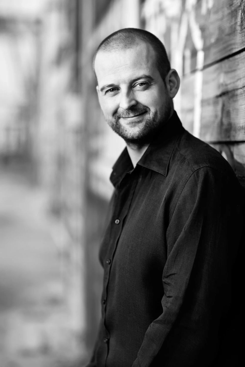 Sort-hvidt portrætfoto af smilende mand med skæg og barbert hoved, lænende sig op ad trævæg i udendørslys.