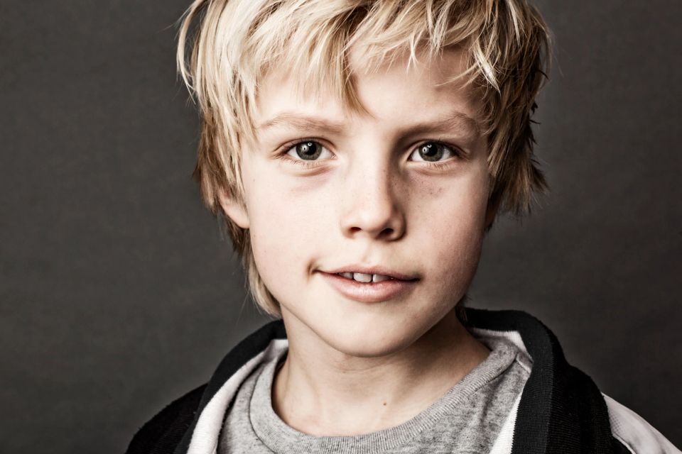 Nært familiefoto af et smilende barn med lyst hår og grønne øjne mod mørk baggrund, naturligt lys. Usyldig glæde.