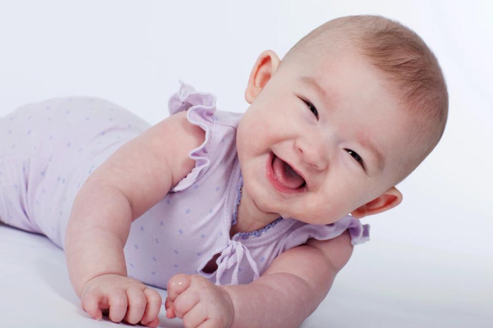 Babyfotografering: Lykkelig baby i lilla sparkedragt med det største smil, liggende på maven mod hvid baggrund.