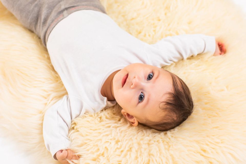 Lille baby med mørkt hår, blåt blik og åben mund liggende på blødt, lyst babylammeskind. Et ægte babyfoto.