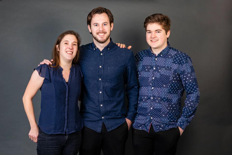 Familiefoto af tre smilende søskende i blåt tøj mod mørk baggrund, som fanger ægte glæde i Virum.