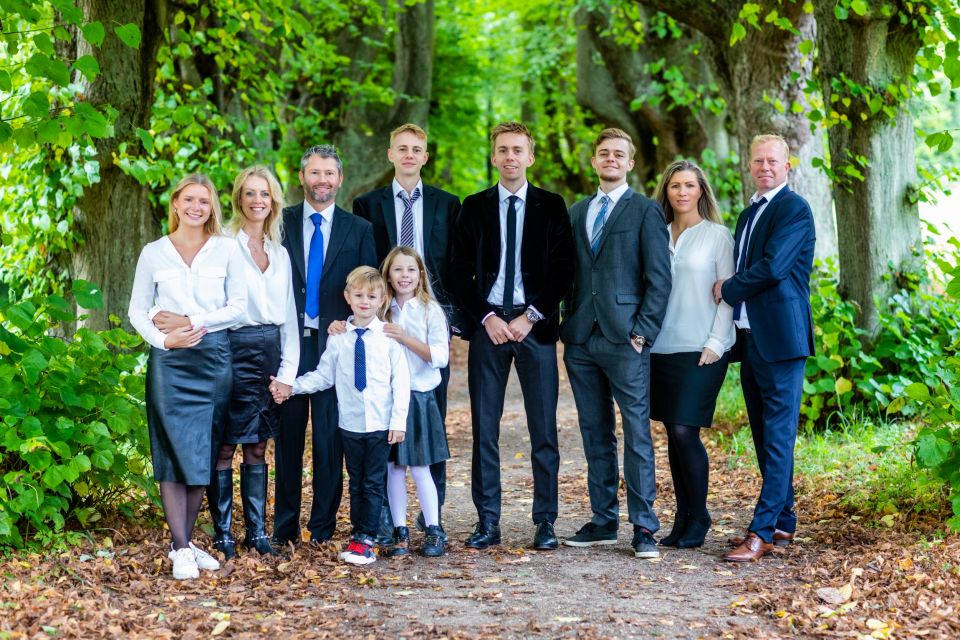 Familiefotografering: Smilende familie på ni personer, voksne og børn, står på en sti i en allé med grønne efterårs blade.