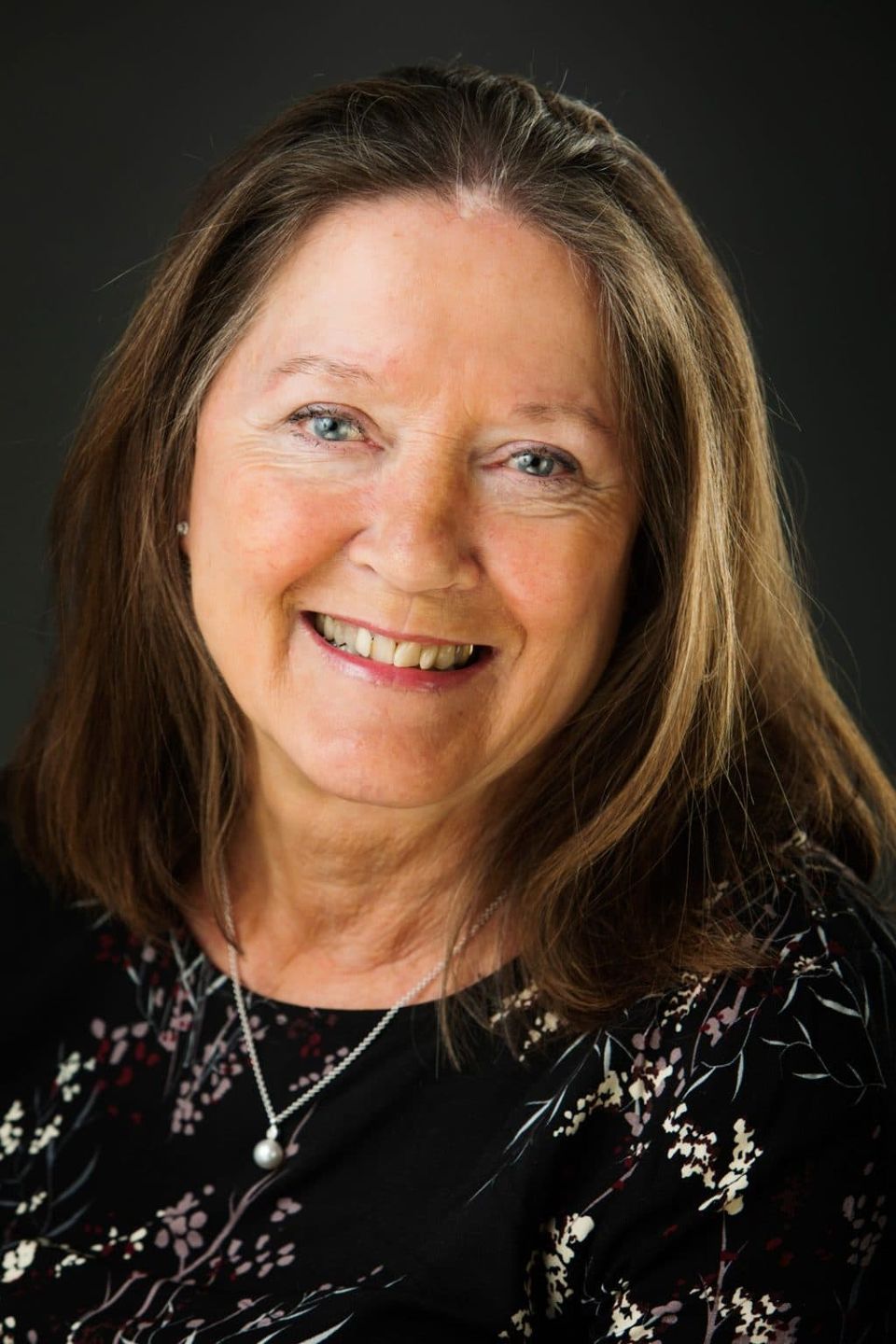 Portrætfoto af smilende kvinde med blåt øje, lyst hår og perlekæde på mørk baggrund i Virum.