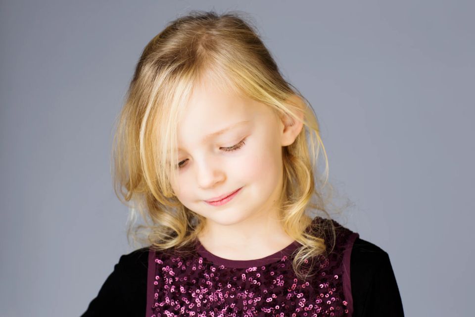Nært børnefoto af smilende pige med lyst hår, der kigger ned. Hun bærer en lilla top med pailletter.
