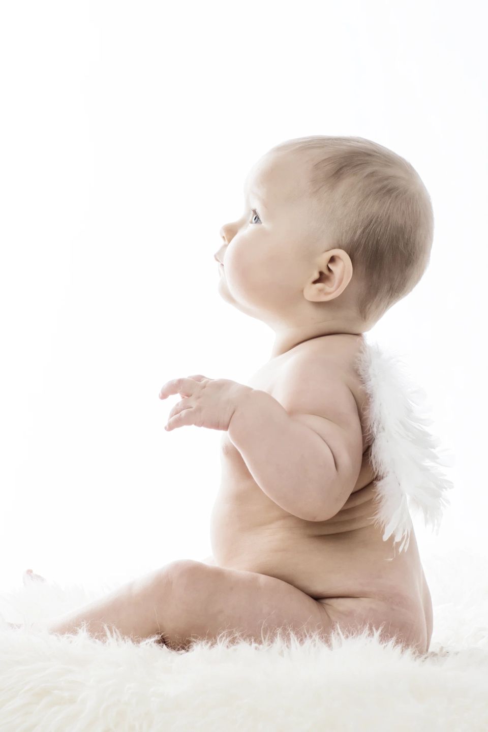 Sødt babyfotografi af spædbarn med englevinger, der sidder på et hvidt lammeskind og kigger op i blødt lys.