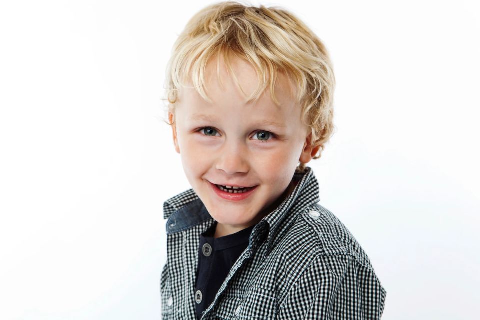 Glad barn med et autentisk smil. Dreng med lyst krøllet hår smiler varmt i blødt fotolys. Børn fotografering.