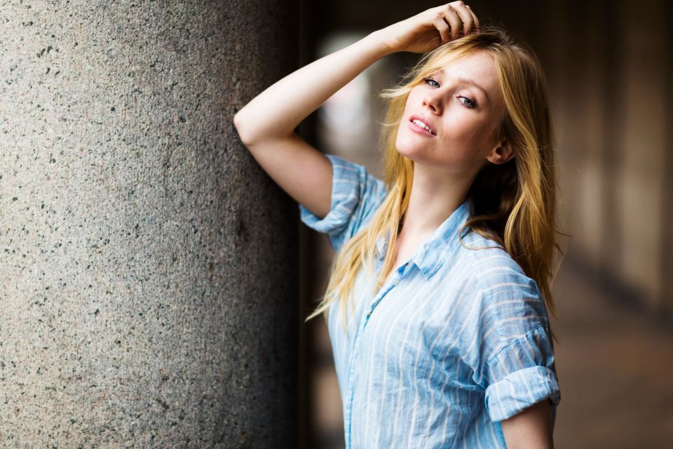 Portrætfoto af smuk, ung kvinde med langt, lyst hår i blå skjorte, der læner sig op ad en søjle i naturligt lys.