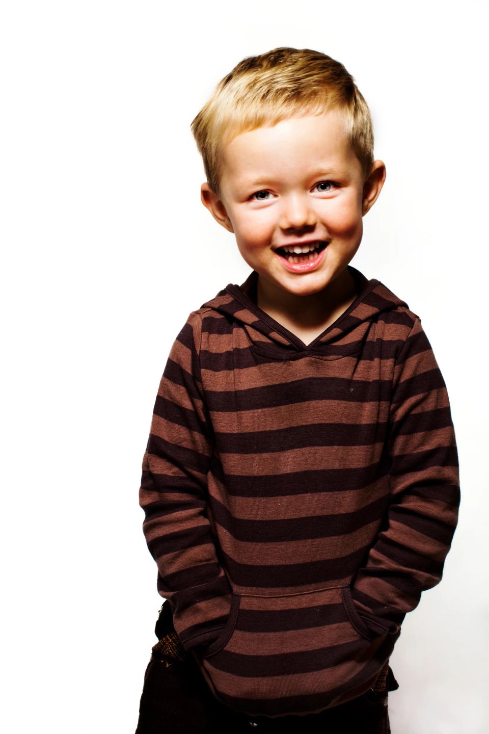 Glad, smilende lyshåret dreng i stribet brun hættetrøje, med hænderne i lommen. Et naturligt børn portrætfoto.