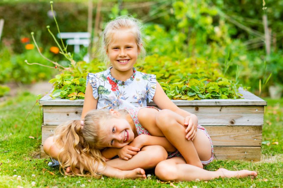Familiefoto af to smilende søstre i en have. Den ene i en blomstret kjole, den anden liggende afslappet på græsset.