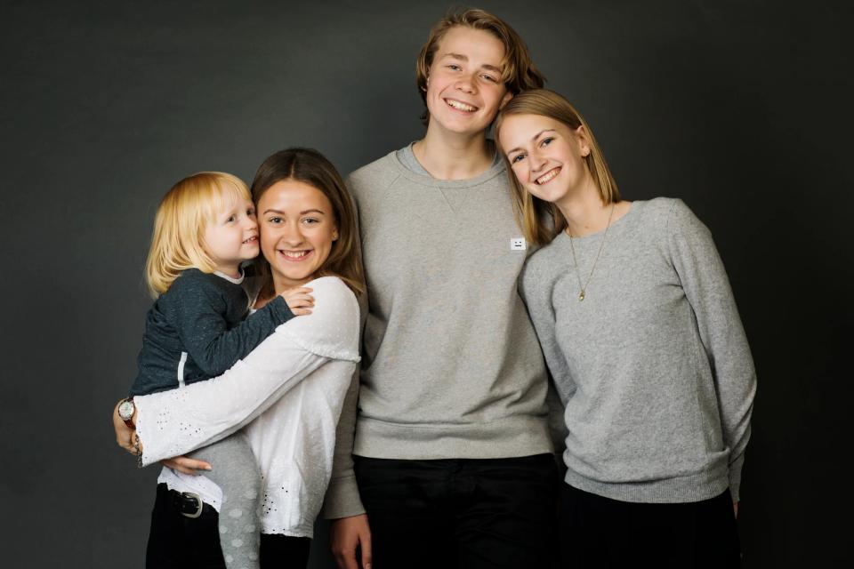 Naturligt familieportræt af fire smilende børn, heraf den yngste på armen, med mørk baggrund. Et skønt børnefoto.