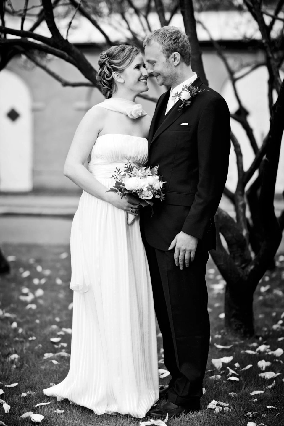 Sort/hvid bryllupsfoto af smilende par, der kigger kærligt på hinanden under nøgne træer. Romantisk stemning.