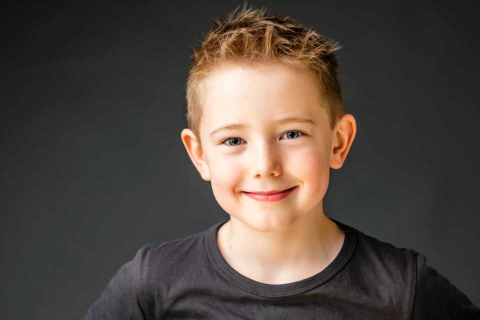 Nærbillede af et smilende barn med blond strithår og blå øjne. Baggrunden er enkel og mørkegrå. Studio familiefoto.