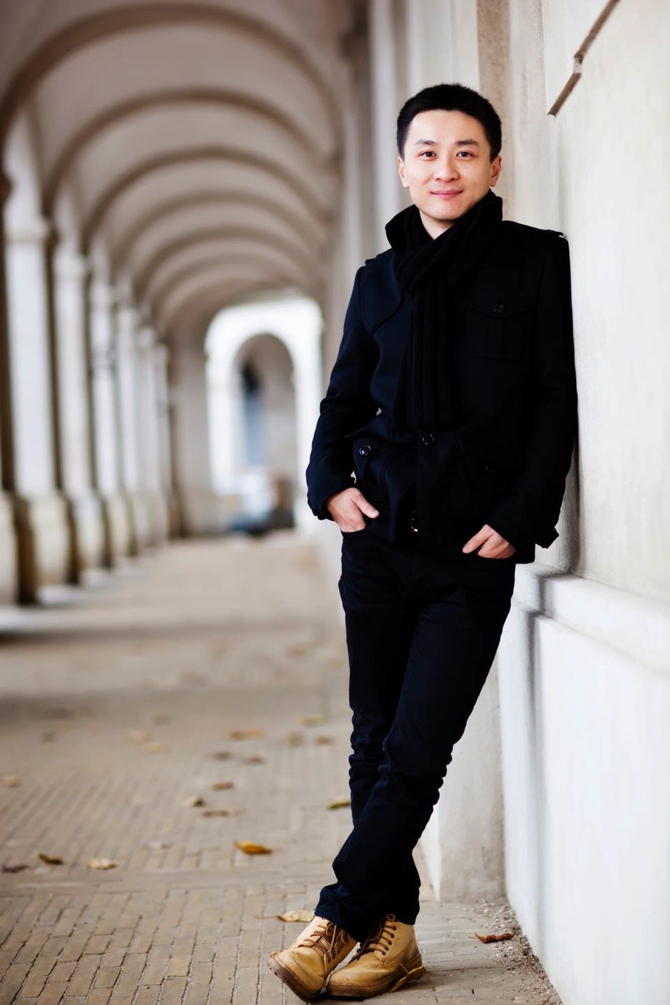 Portrætfoto af mand i sort jakke og halstørklæde, som smiler venligt og står lænet op ad en væg i en søjlegang.