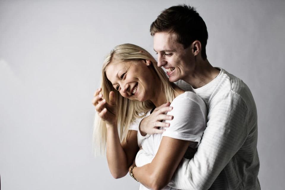 Glad parfotografering: Et portrætfoto af et ungt par, hvor manden krammer kvinden bagfra, begge smiler ægte og hjerteligt.