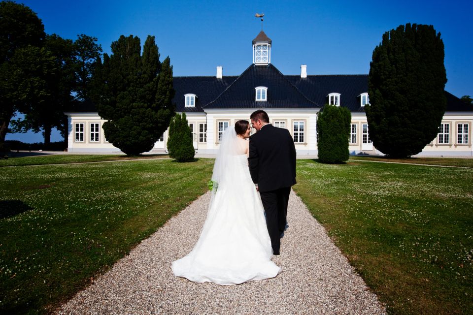Bryllupsfotografi: Brudepar hånd i hånd går væk fra kameraet på en grussti mod et hvidt gods under blå himmel.