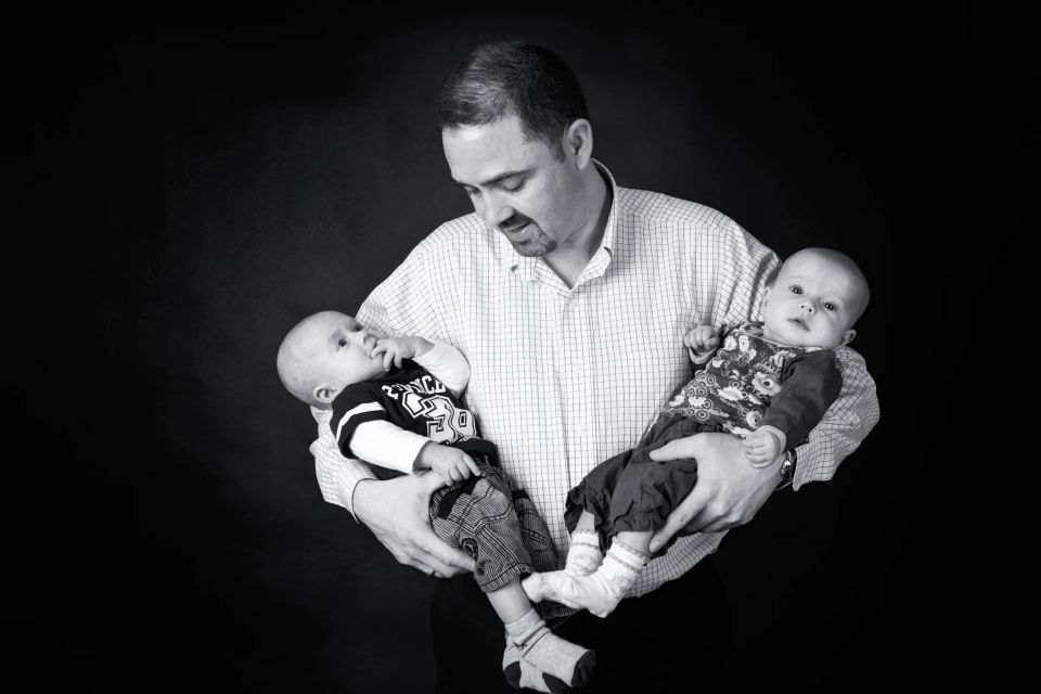 Sort-hvid familiefoto af far der kigger kærligt på tvillinger. Et autentisk portræt med fokus på relationen.