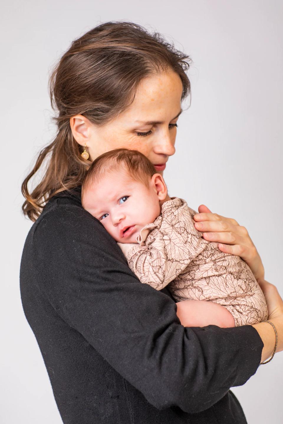 Sort/hvid babyfotografering: Mor holder sit spædbarn tæt, babyen kigger direkte mod kameraet i blødt lys.