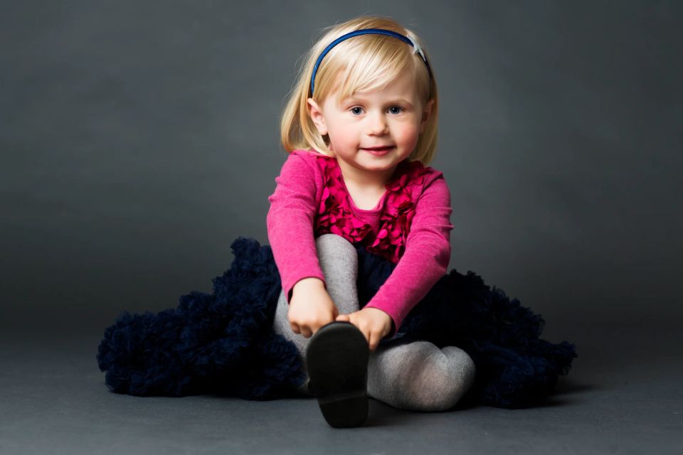 Børnefotografering af blond smilende pige i lyserød bluse og mørkeblåt skørt, der sidder på gulvet.