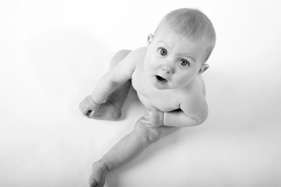 Sort-hvid babyfotografering med nysgerrig baby liggende på maven, kigger op med store øjne og åben mund.