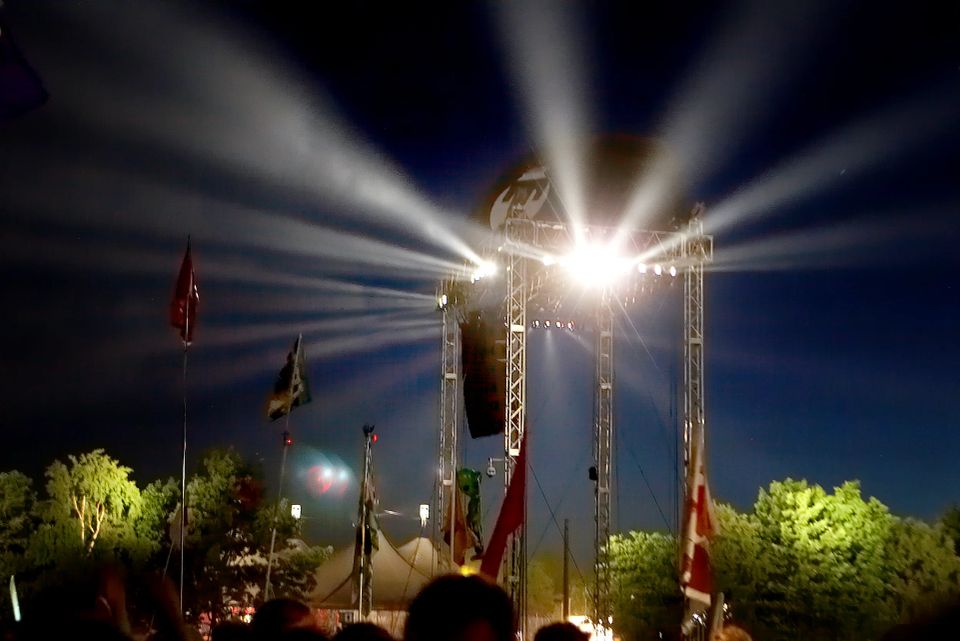 Nattefoto af et Roskilde Festival lysshow med kraftige stråler fra scenen, der lyser op mod den mørke himmel over publikum.