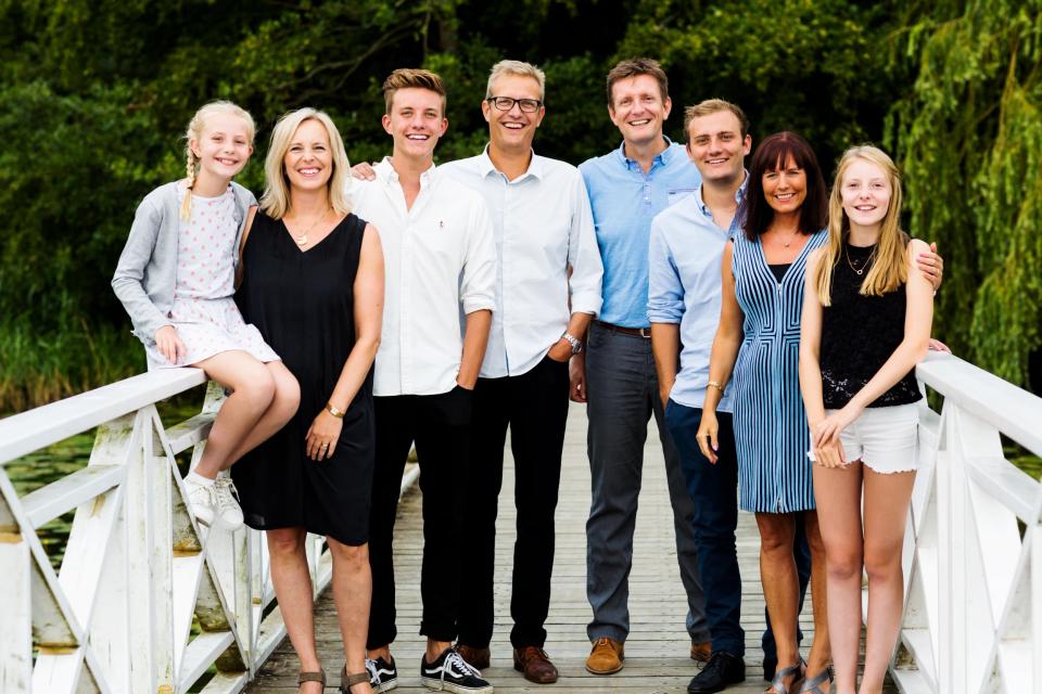 Familiefoto af syv smilende personer i forskellige aldre på en hvid træbro i Virum, med grønne træer i baggrunden.