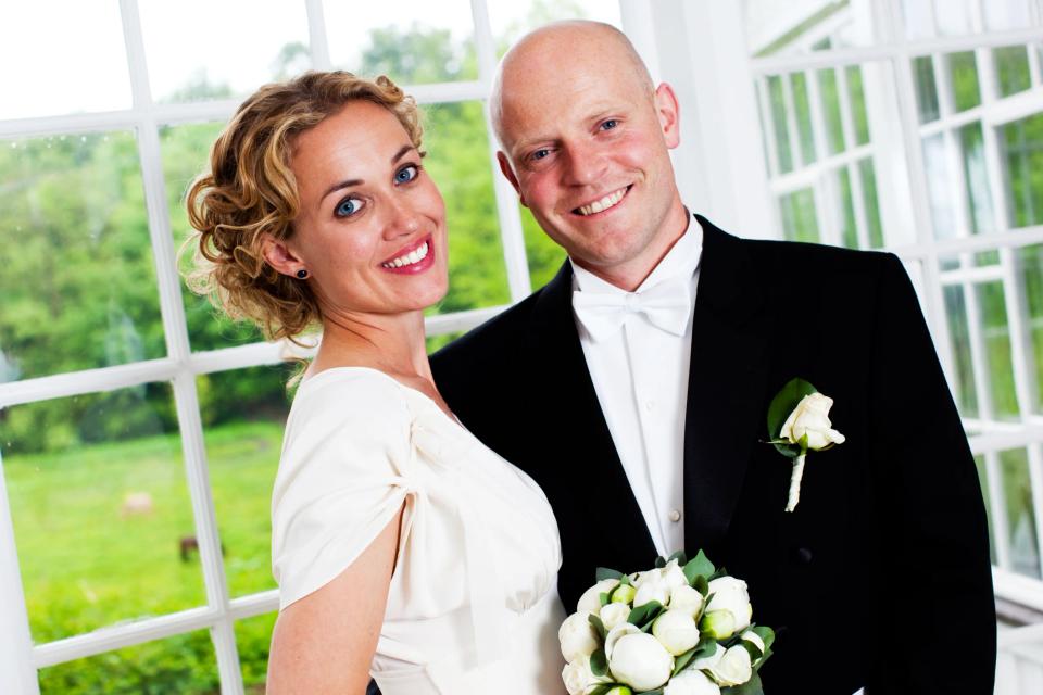 Bryllupsfoto af smilende par med brudebuket, stående elegant foran et vindue med grønne træer i baggrunden.